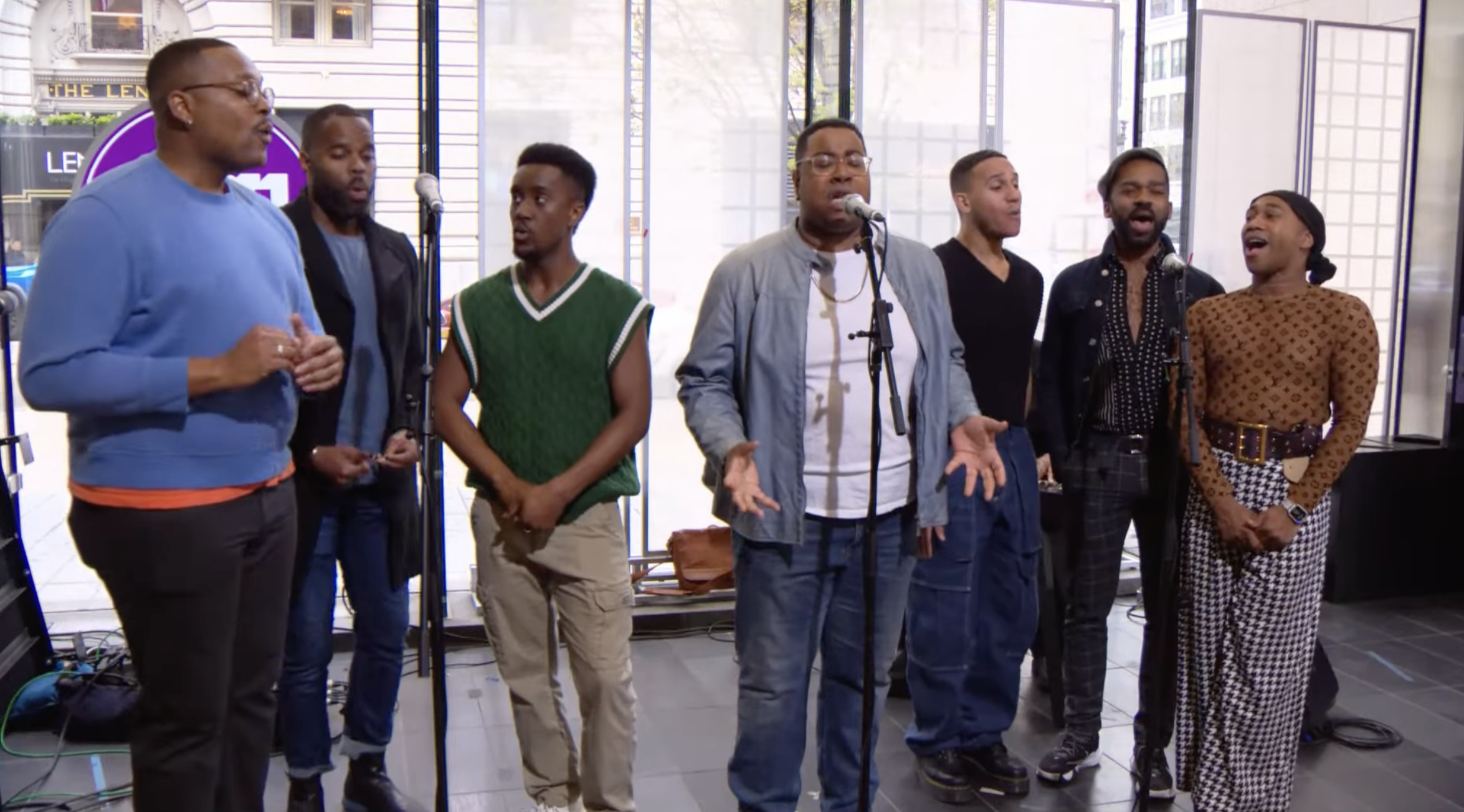 A group of eight people singing together in a living room with large windows and a city street view outside. They are standing in front of microphones, with some using their hands to express emotion.