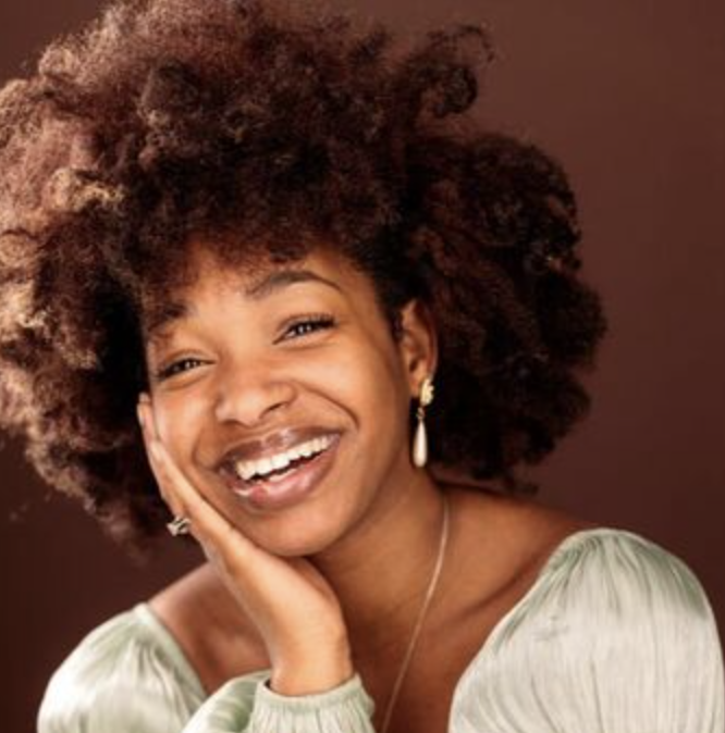 A young woman with natural curly hair smiling and resting her face on her hand, wearing earrings and a light-colored top, against a brown background.