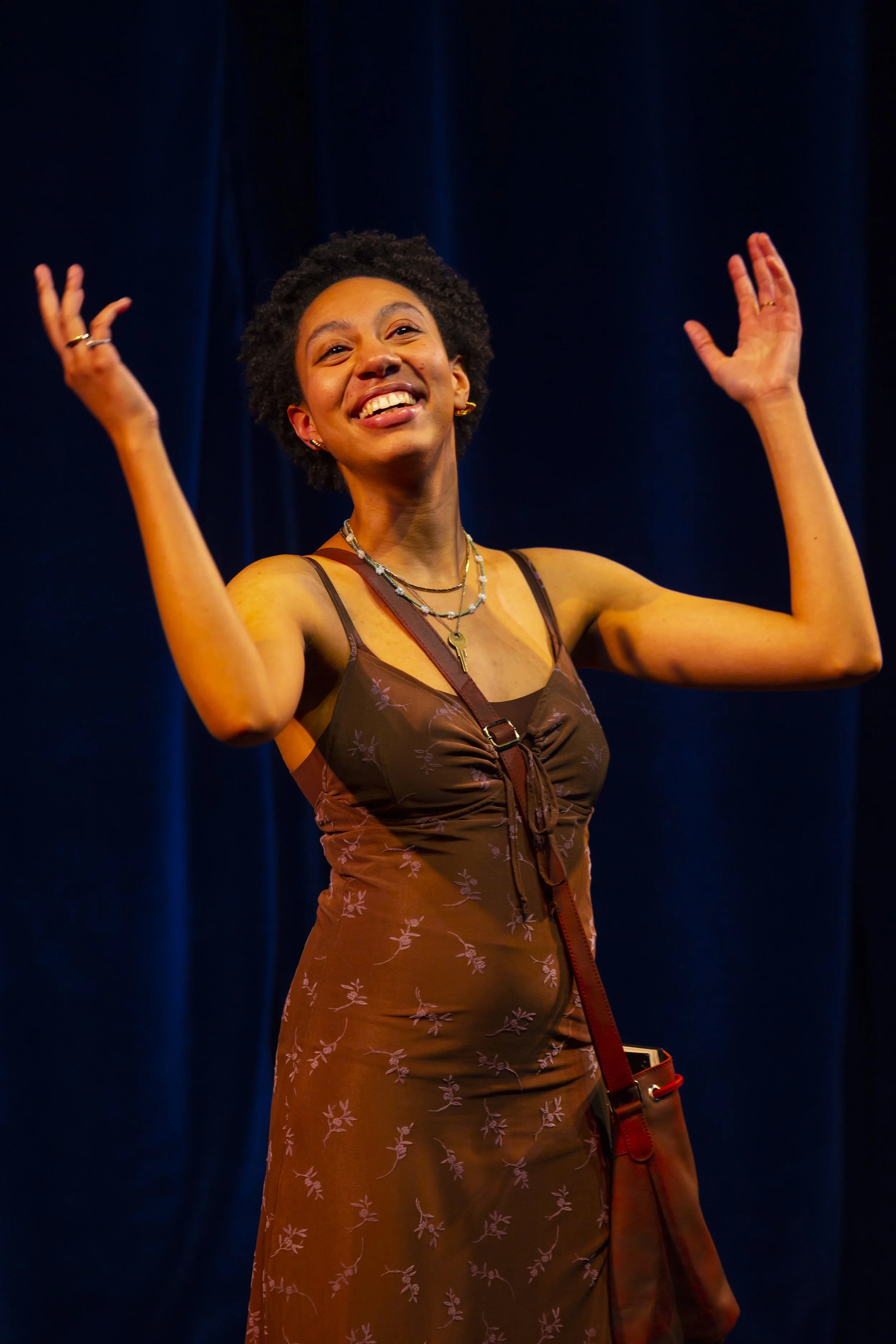 A woman with short curly black hair and earrings, smiling with her hands raised, wearing a brown dress with a floral pattern and layered necklaces, standing against a dark blue curtain background.