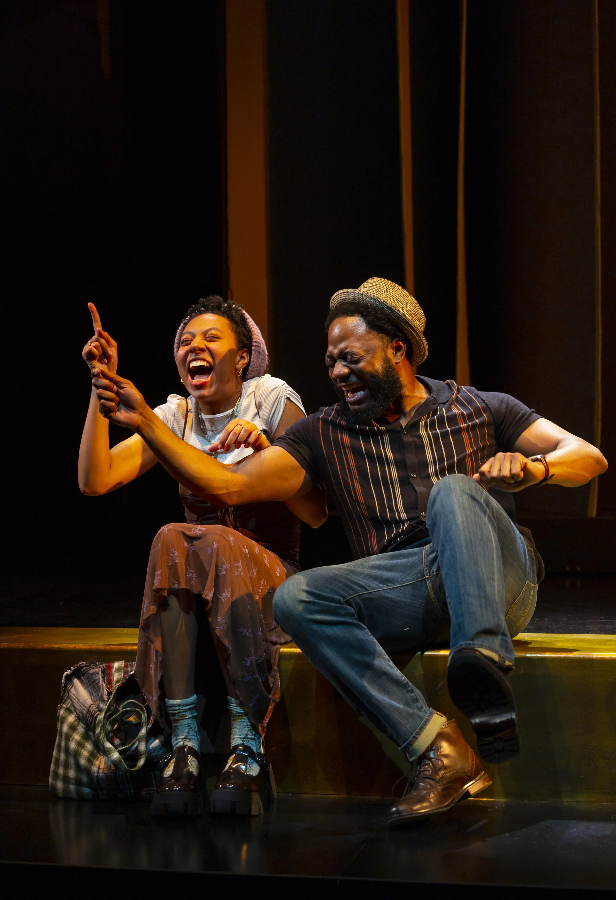 Two performers sit on stage, laughing and enjoying a moment together. The woman on the left raises her index finger while the man on the right leans forward, showing expressive faces. The stage background is dark with vertical wooden panels.