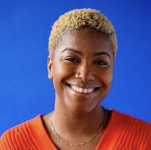Close-up portrait of a smiling woman with short blonde hair wearing a red top and gold necklace against a blue background.