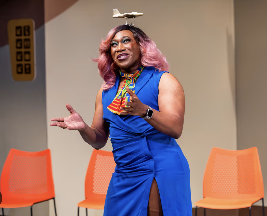 A woman with pink hair and a blue dress standing on a stage, speaking and gesturing with her hands. She has a small model airplane balanced on her head. Four orange chairs are visible behind her.