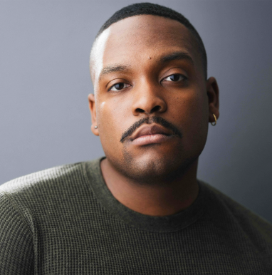 Close-up portrait of a young man with short hair, a mustache, and goatee, wearing a dark green shirt and a small hoop earring on his left ear, against a plain gray background.