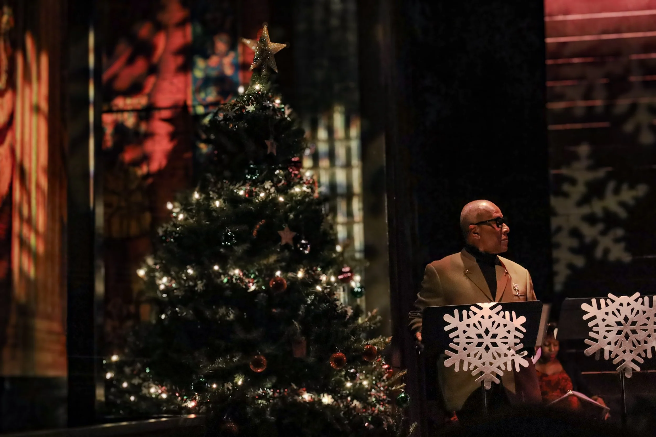 A decorated Christmas tree with ornaments and a star on top, beside a man in a formal suit and glasses playing a piano with snowflake decorations.