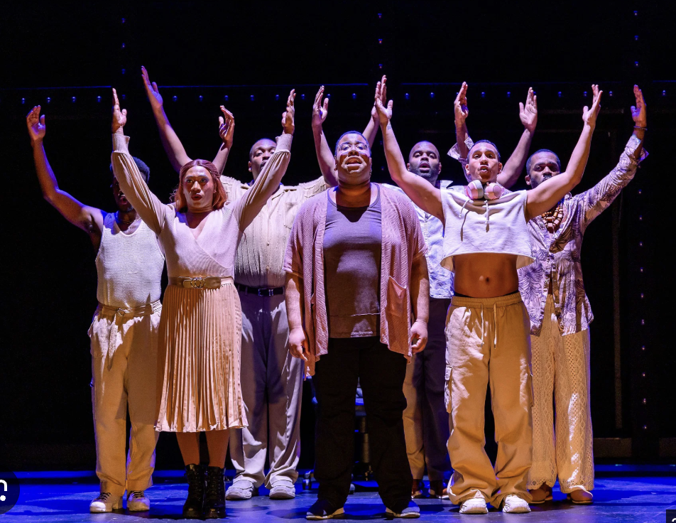 A diverse group of seven performers on stage, standing with arms raised, singing or acting, under stage lighting.