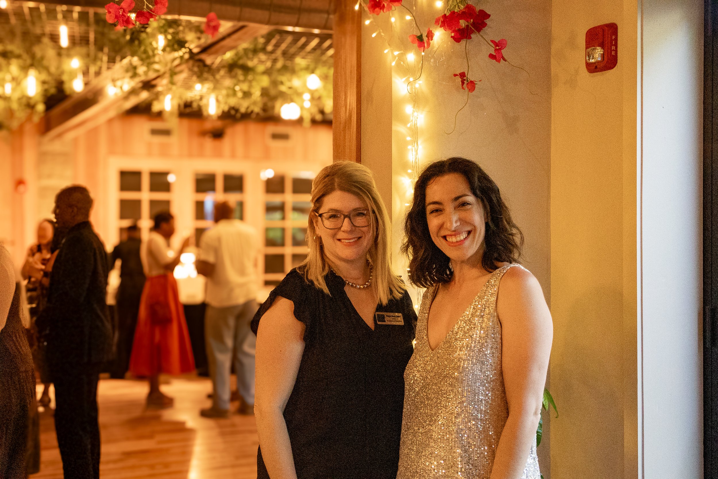 Two women smiling and standing close together at an indoor event. One woman wears glasses and a black dress with a pearl necklace, and the other wears a sparkling gold dress. The background shows warm lighting, string lights, and other guests minglin
