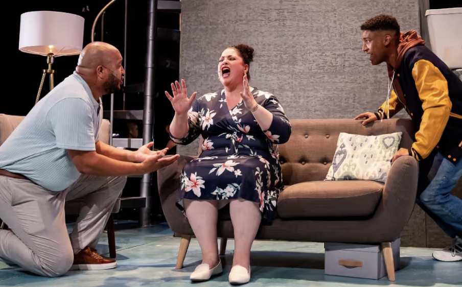 Three people having a heated argument in a living room, with one woman sitting on a sofa and two men standing and kneeling, all appearing to be angry.