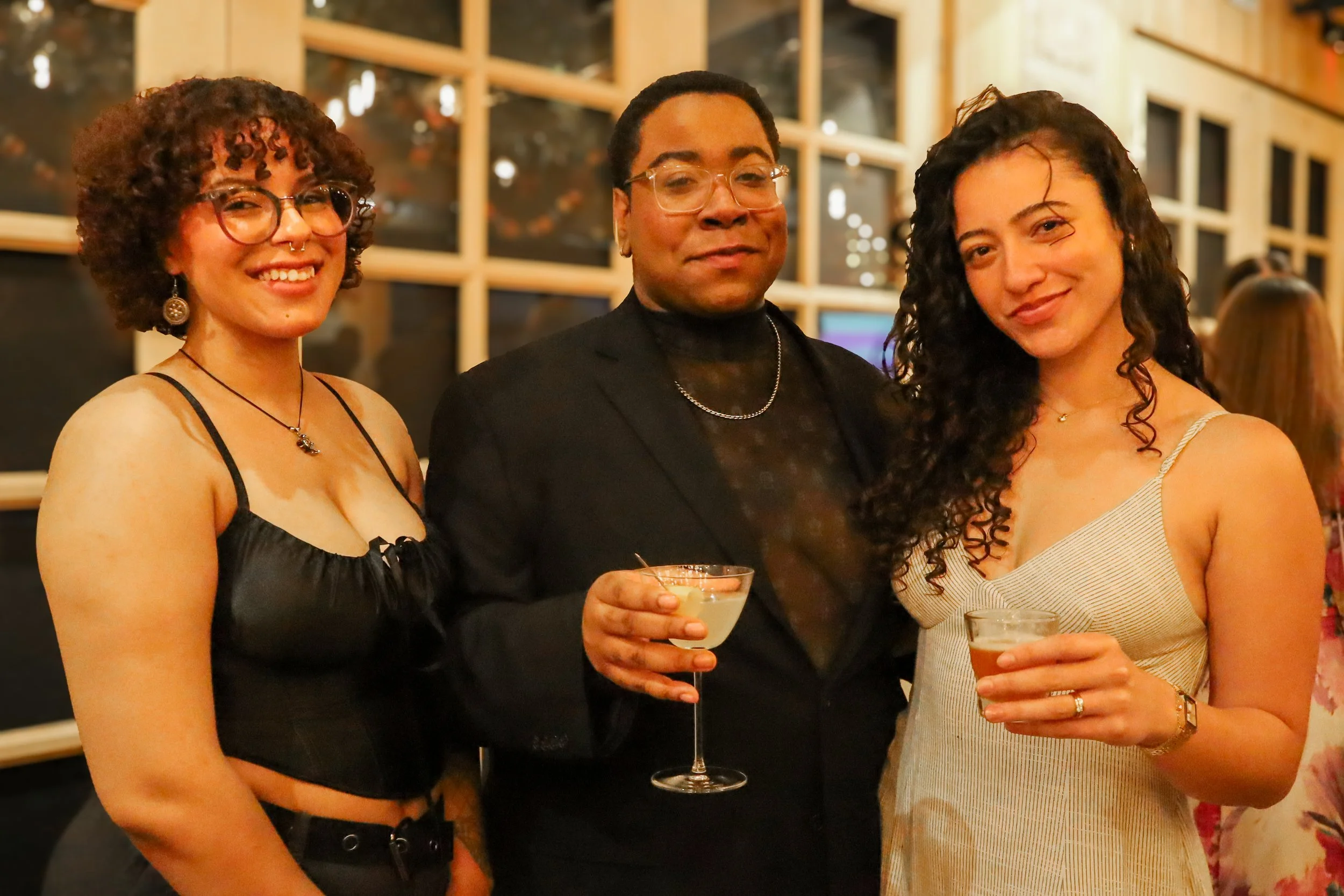 Three women and one man enjoying drinks at a social event, smiling for the camera, standing in front of a decorative background with wooden shelves, in a warmly lit indoor setting.