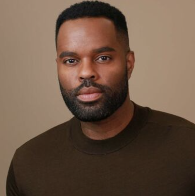 Portrait of a man with short black hair and a beard, wearing a dark brown shirt, against a neutral background.