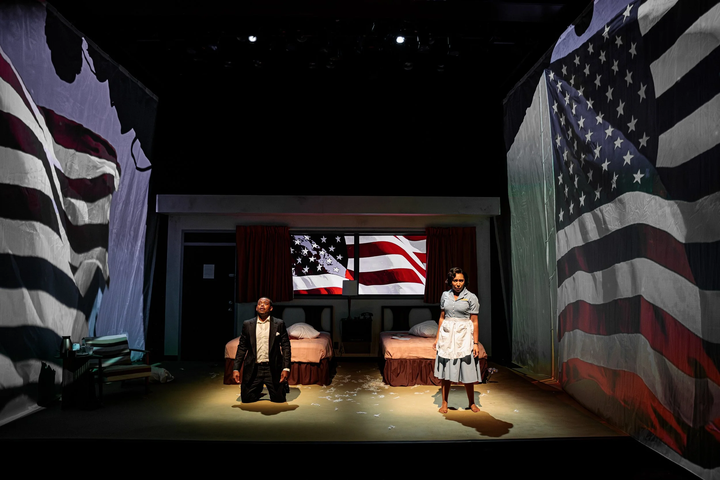 Two actors on a stage with American flags hanging on the sides and in the background. One actor is kneeling and the other standing, both in period costumes, with a dimly lit scene.