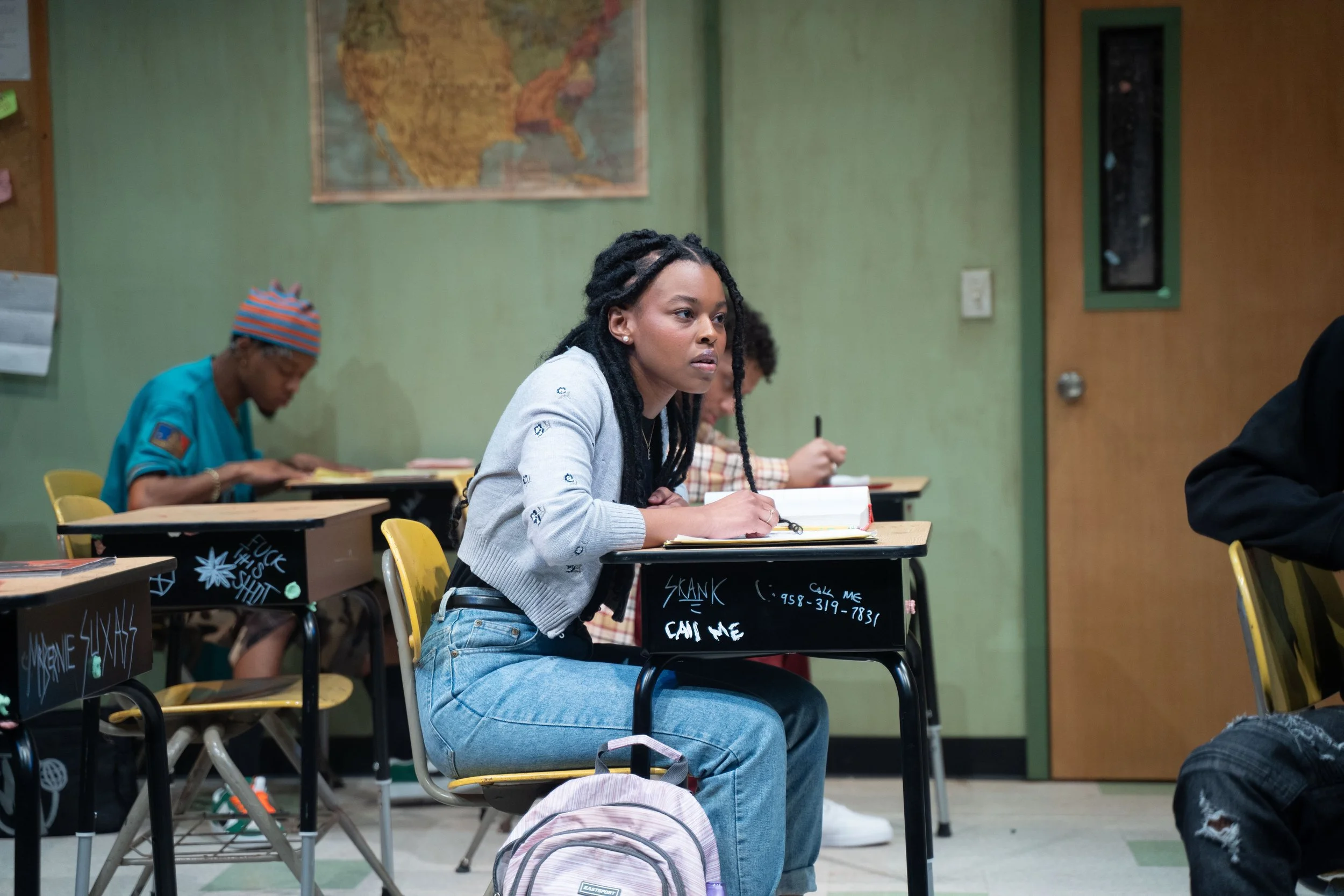 A classroom scene with a young woman with long dreadlocks sitting at a desk, looking to the side, with notebooks and a backpack nearby. Other students are in the background, including an older woman with a striped beanie. The classroom has green walls, a world map, and various bulletin boards.