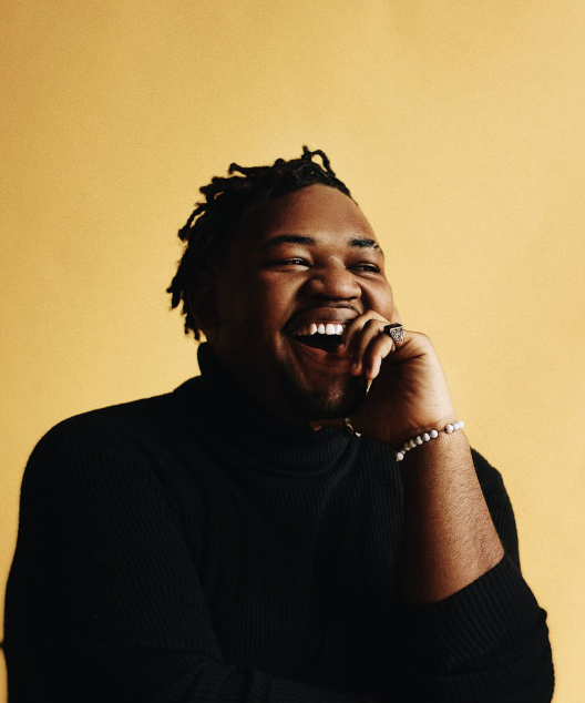 A smiling man with dreadlocks wearing a black turtleneck and jewelry, against a yellow background.