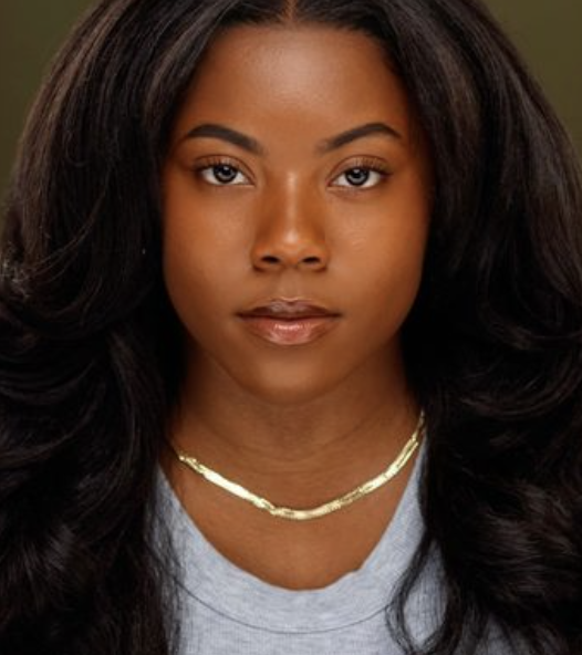 Close-up portrait of a young woman with long, dark, wavy hair, wearing a light gray top and a gold necklace.