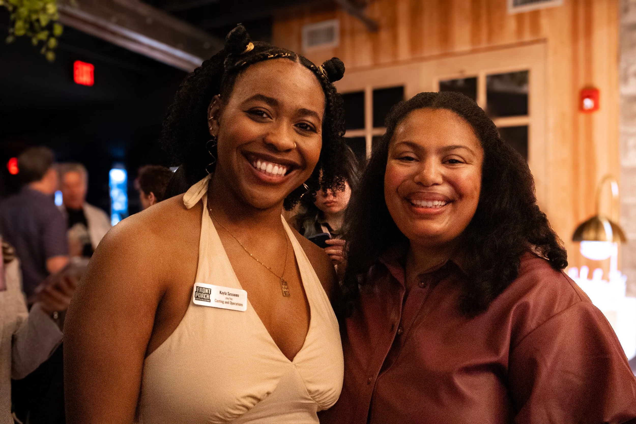 Two women smiling, one with a name tag, in an indoor setting with wooden walls and other people in the background.