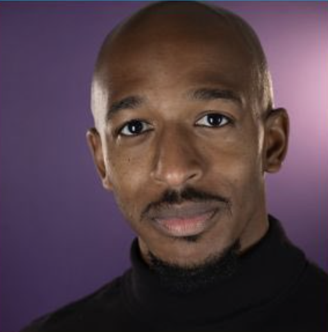 Close-up portrait of a man with a shaved head, mustache, and goatee, wearing a black turtleneck against a purple background.