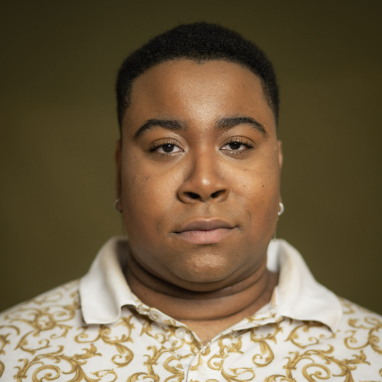 Close-up of a woman with short hair, wearing a white shirt with gold patterns, looking directly at the camera with a neutral expression.