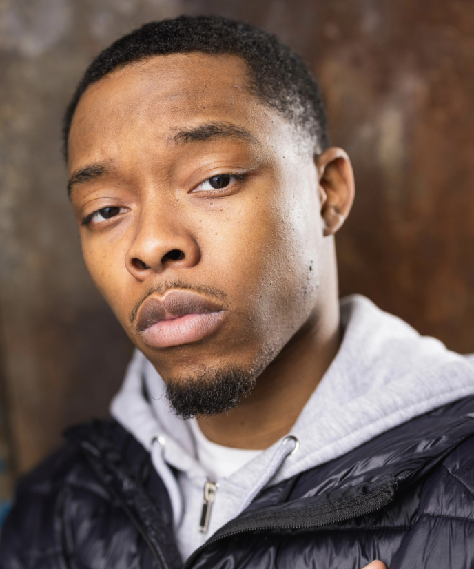 Close-up portrait of a young man with short black hair, facial hair, wearing a gray hoodie and black jacket, against a blurred brown background.