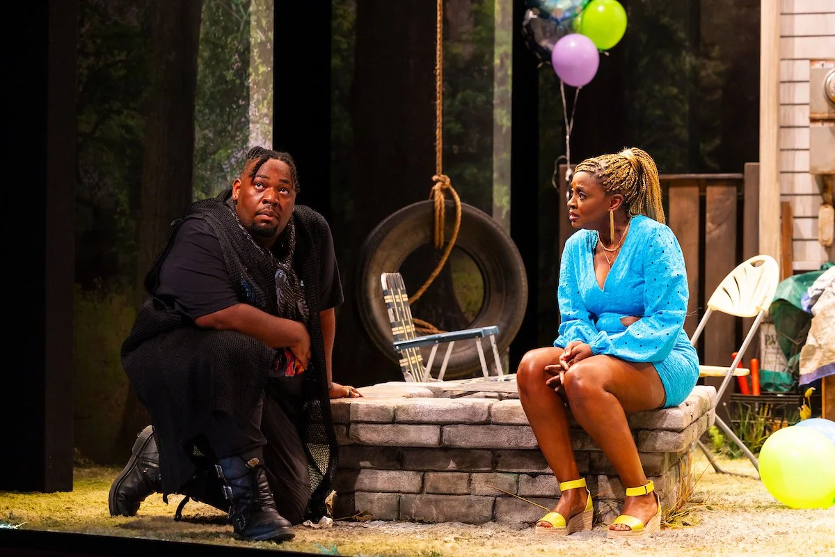 Two people sitting and kneeling next to a brick well, with a tire swing hanging behind them and balloons in the background.