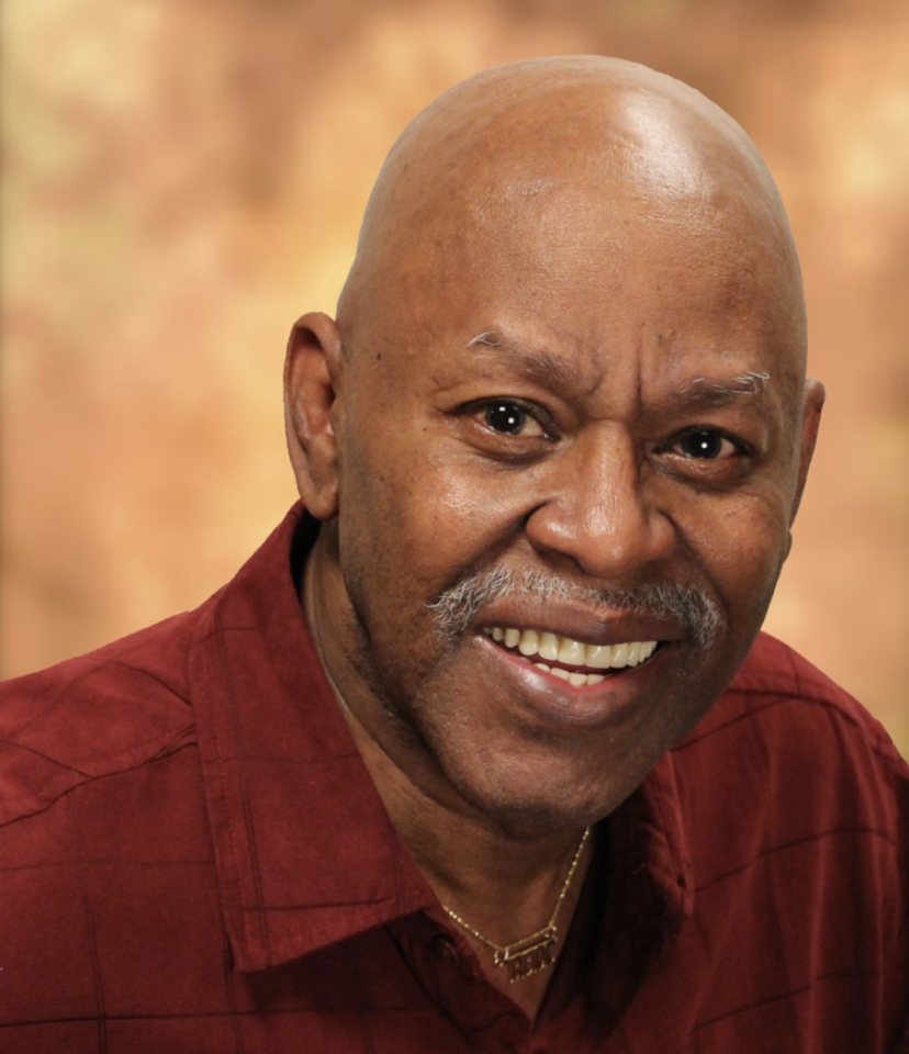 A smiling man with dark skin, bald head, and gray beard wearing a maroon shirt, against a blurred warm background.
