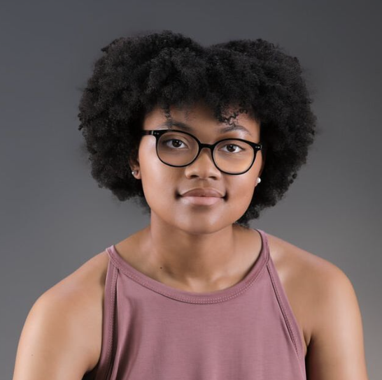 Portrait of a young woman with dark curly hair, glasses, and earrings, wearing a sleeveless mauve top against a gray background.