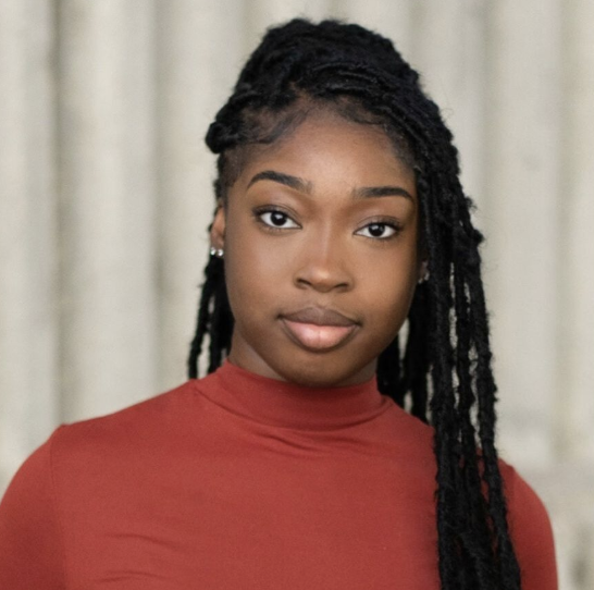 A young African American woman with long braided hair wearing a red turtleneck, looking at the camera with a neutral expression, standing in front of a beige curtain.