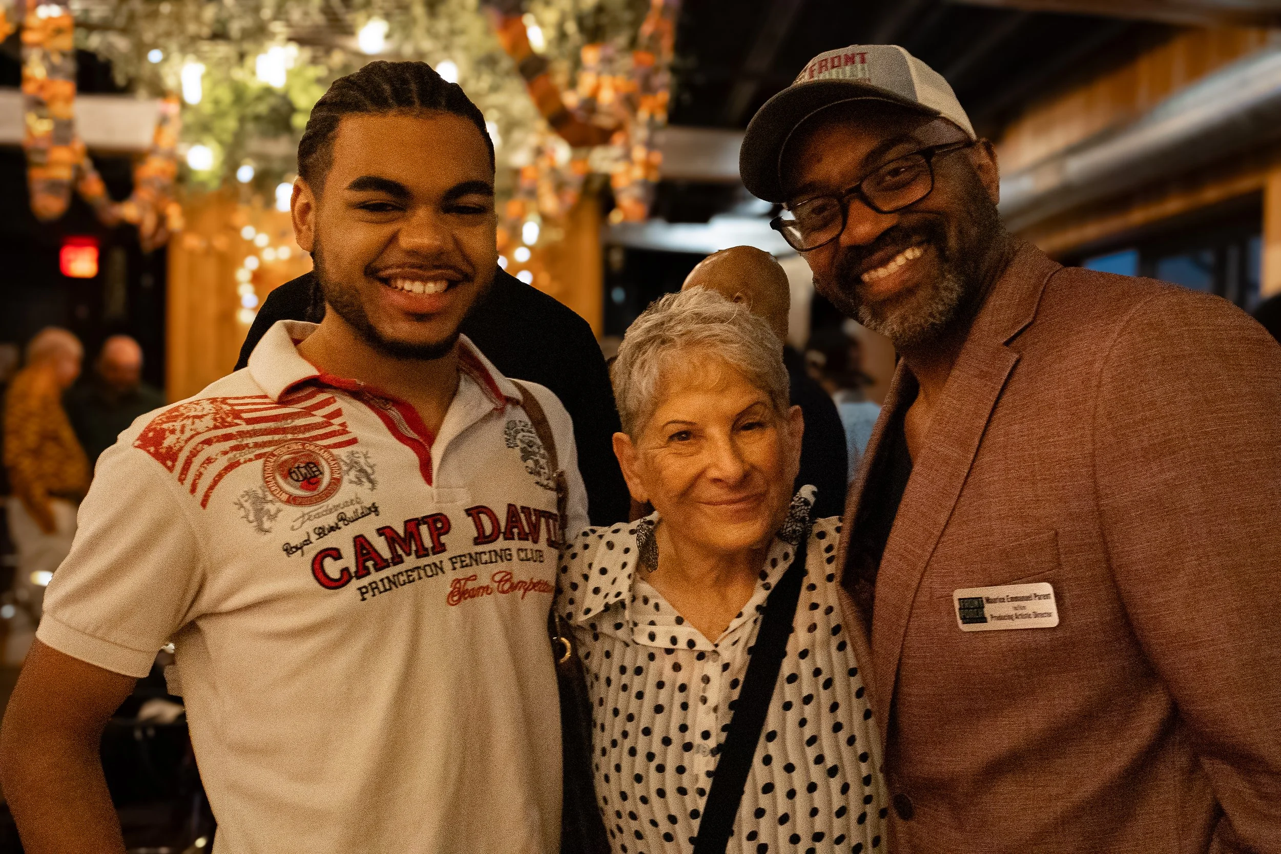 Three people smiling and posing together indoors at a social event, with a warm, decorated background.