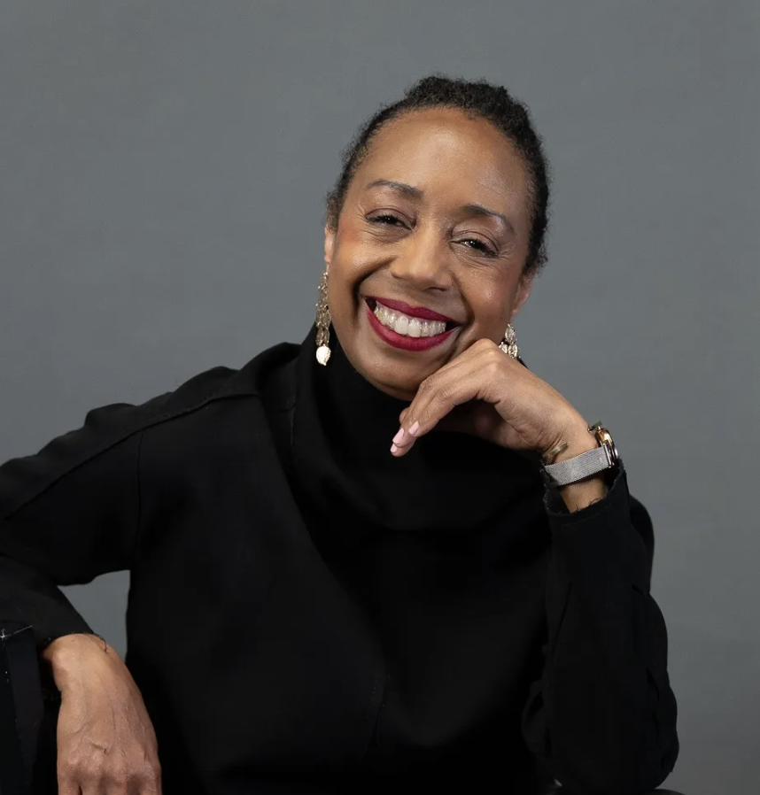 A woman with dark hair, wearing earrings, a watch, and a black turtleneck sweater, posing against a gray background, smiling and resting her chin on her hand.