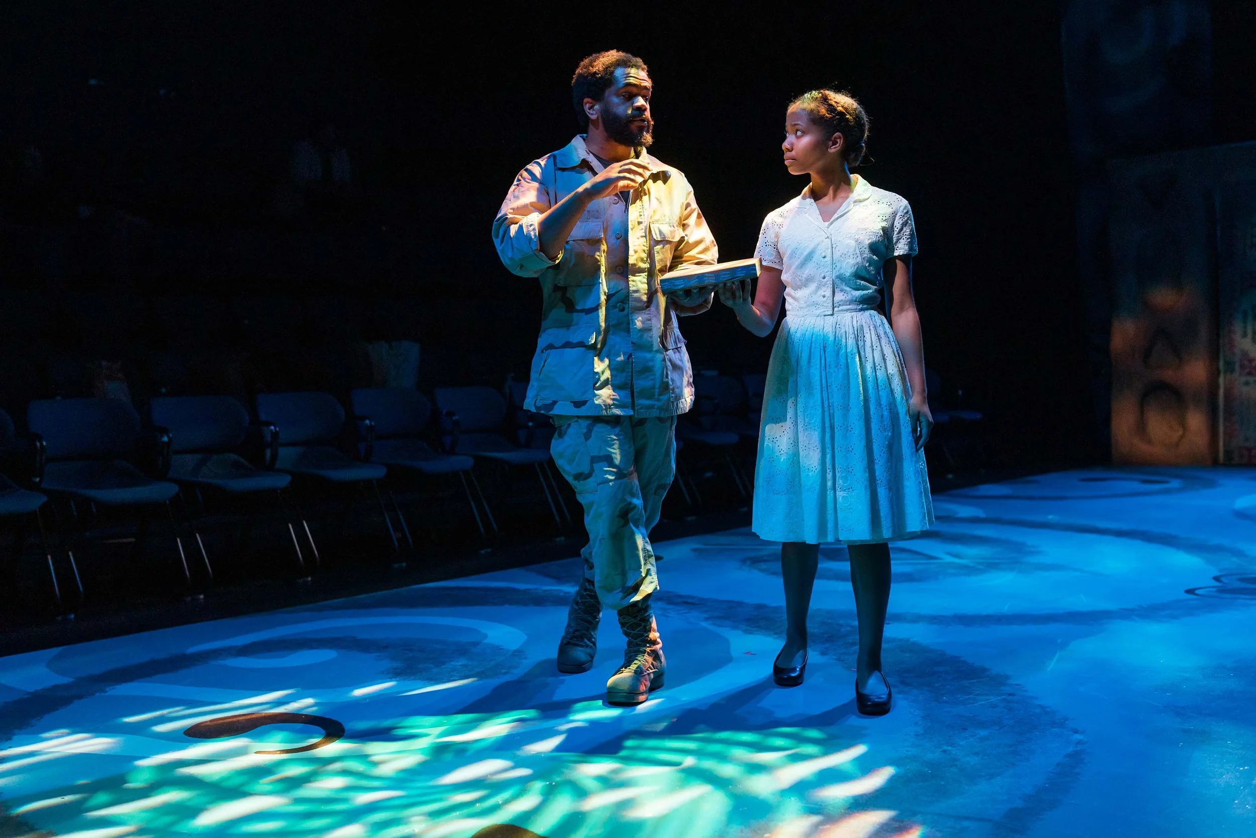 Two actors on stage, a man in military-style clothing and a woman in a white dress, engaged in a scene with dramatic lighting.