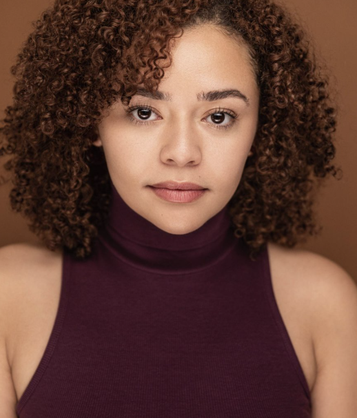 A young woman with curly hair wearing a sleeveless purple turtleneck, looking directly at the camera with a neutral expression