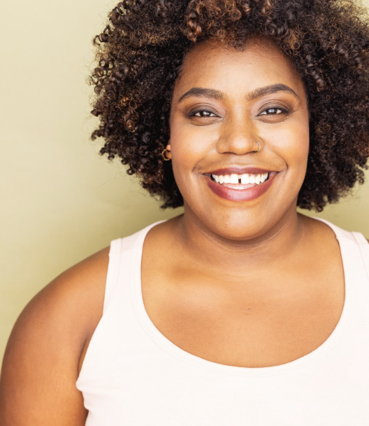 Close-up of a smiling woman with curly hair, wearing a white tank top and a nose ring, standing against a pale green background.