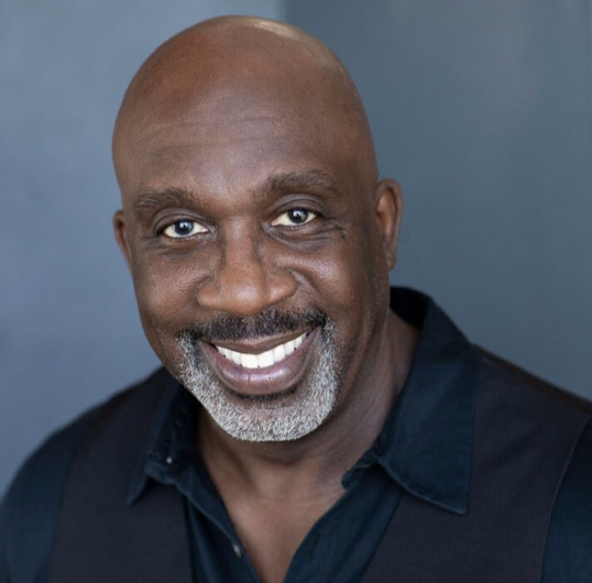 Close-up portrait of a smiling bald African American man with a gray beard, wearing a dark collared shirt, against a muted blue background.