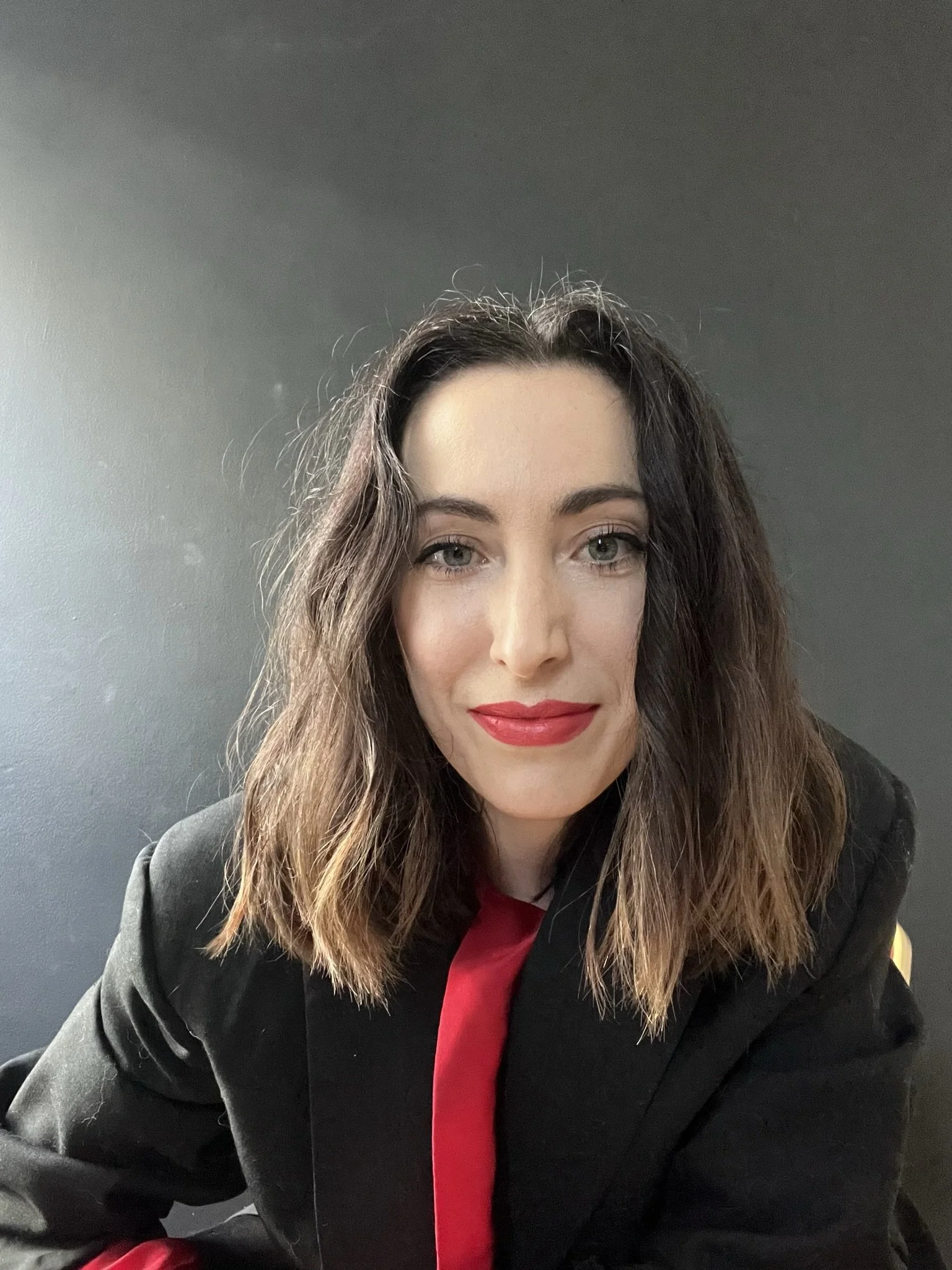 A woman with long, wavy brown hair and light skin, wearing a black blazer, a red shirt, and bright red lipstick, sitting against a plain gray background.