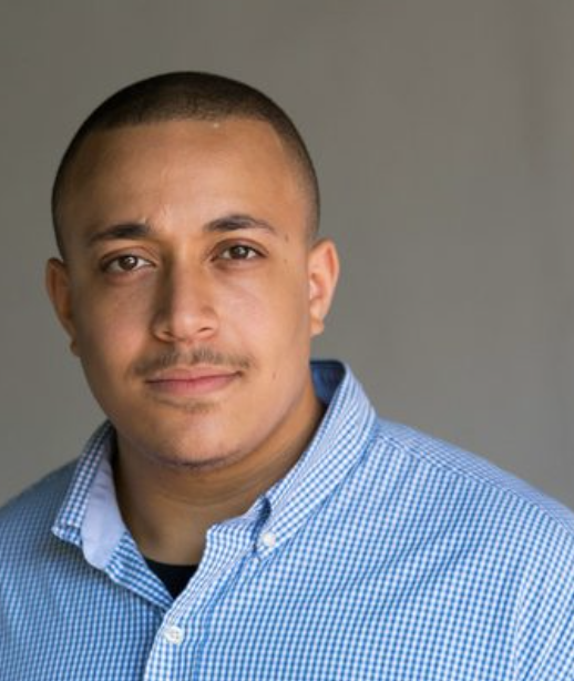 A young man with short hair and light brown skin, wearing a blue checkered button-down shirt, posing against a plain gray background.