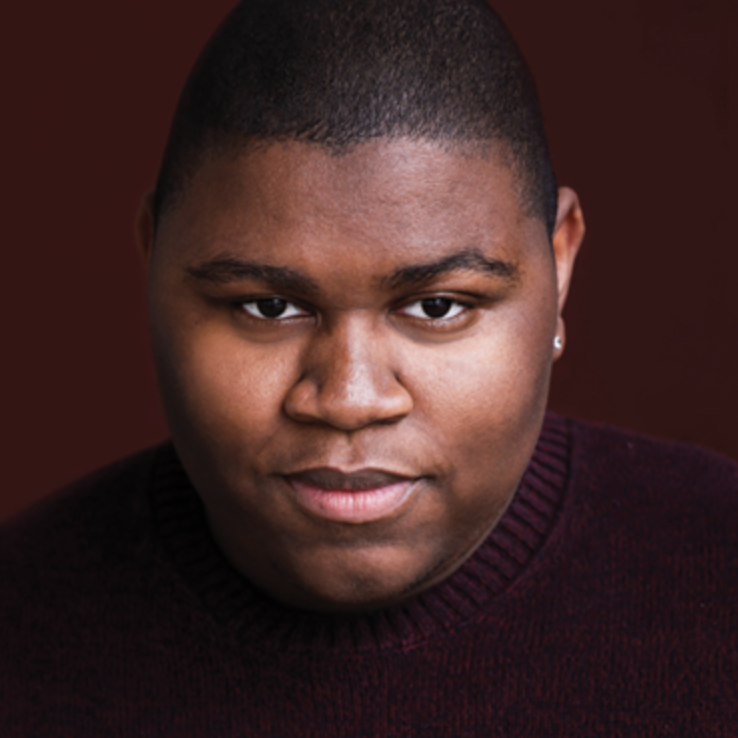 Close-up portrait of a young man with short hair, wearing a dark maroon sweater, looking directly at the camera with a slight smile, against a dark background.