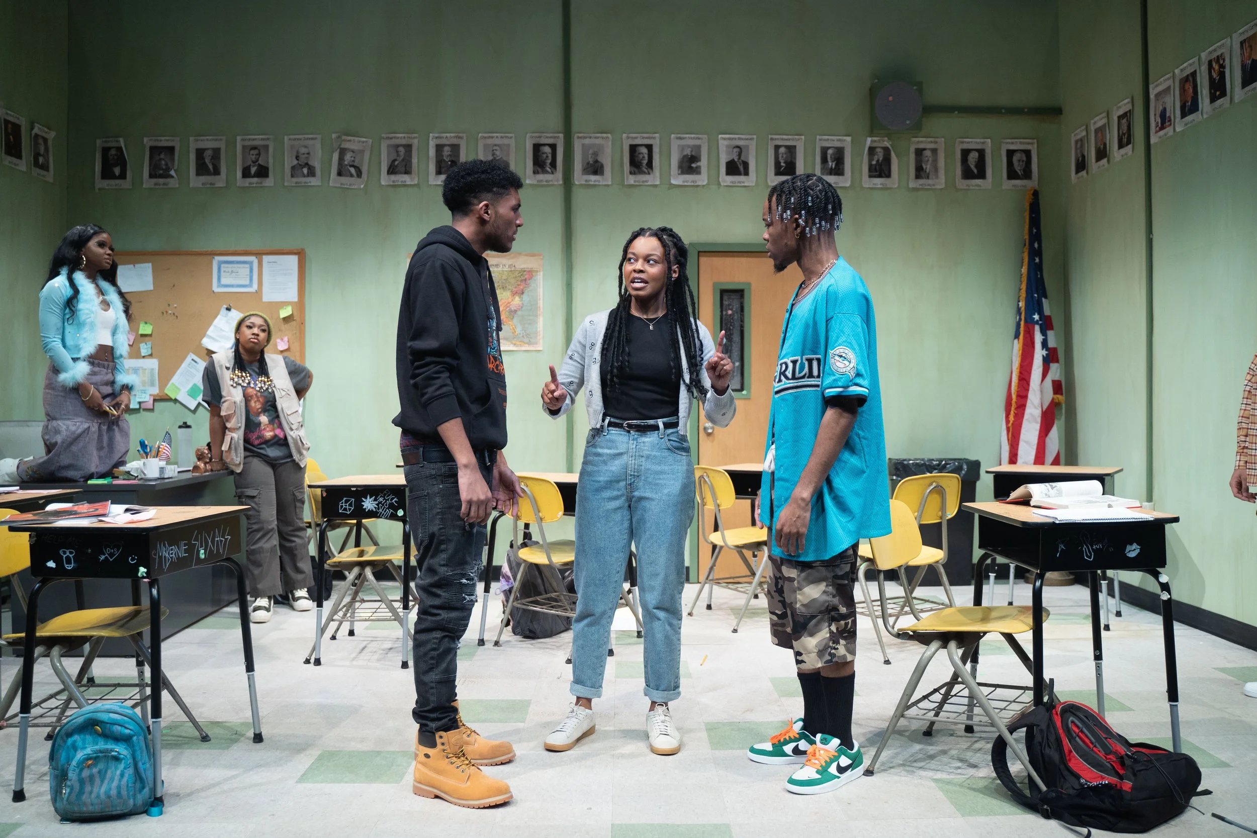 Four young people stand in a classroom, engaged in conversation. Two are facing each other in the center, one male wearing a black hoodie and one female with long braided hair. The third person, also male with braided hair, is to the right and facing them. The fourth person, a female with long dark hair, stands behind them, observing. Classroom desks and chairs are visible, along with backpacks, books, and classroom decorations.