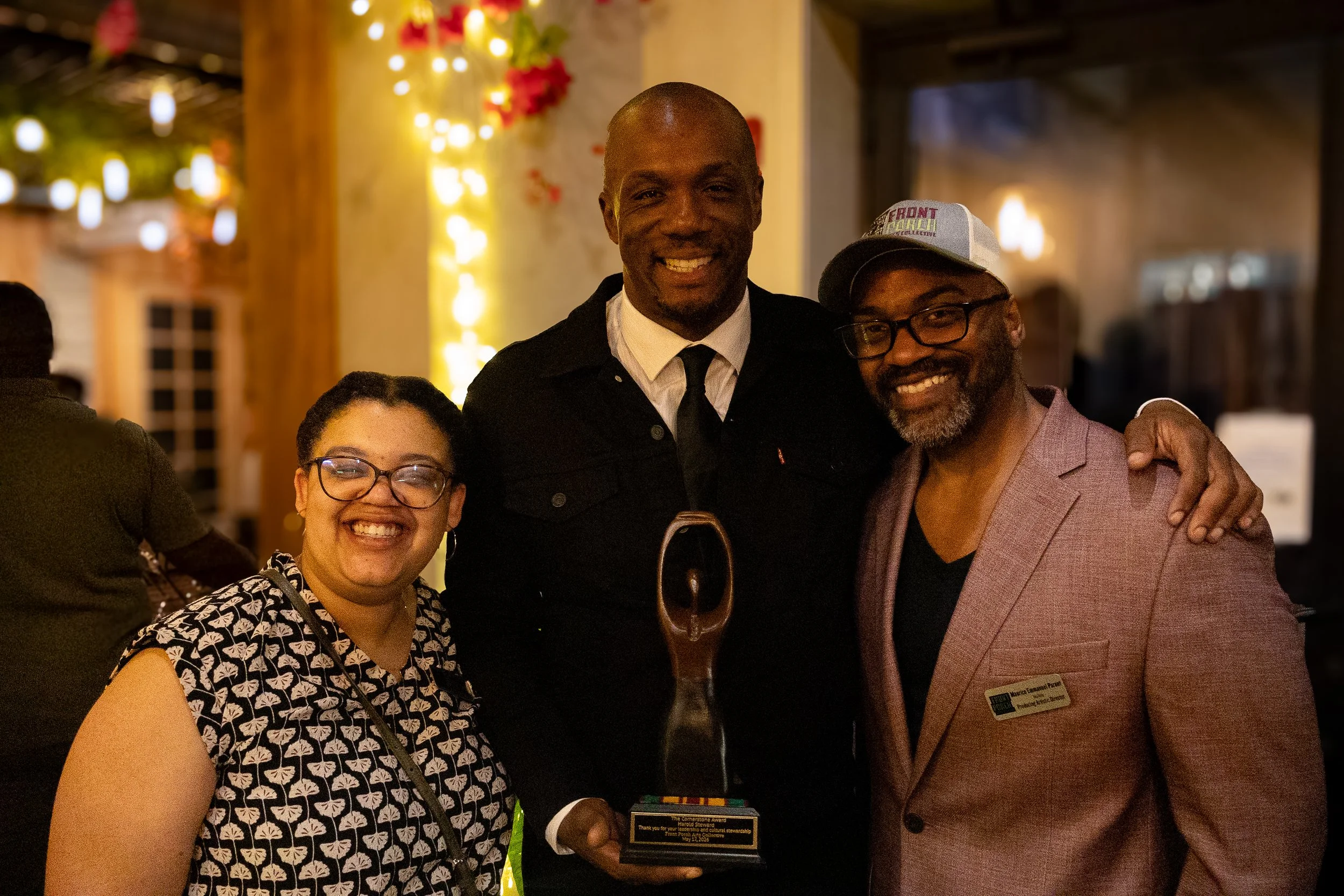 Three smiling people at an event, with a man in the center holding an award plaque. The background has warm lighting and decorative lights.