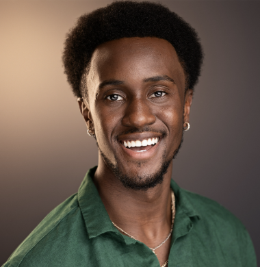 A smiling young African American man with natural hair, wearing a green shirt and a necklace, against a neutral background.