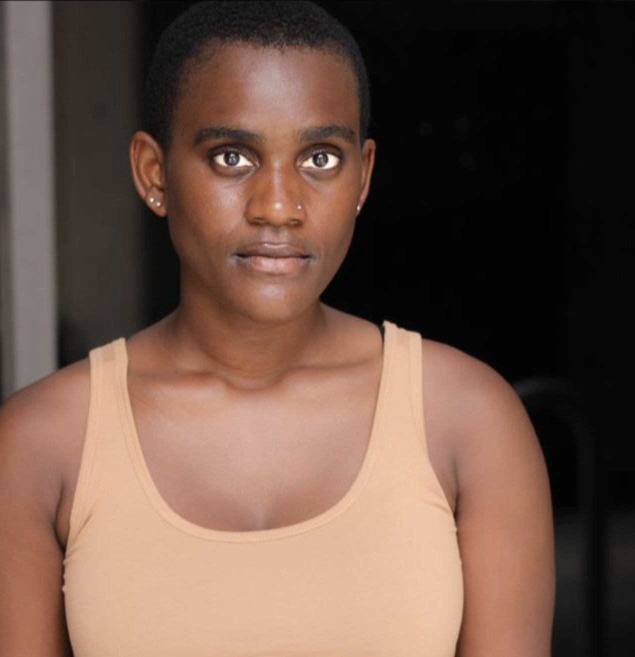 Close-up of a young woman with dark skin and short hair, wearing a beige tank top, looking directly at the camera with a neutral expression.