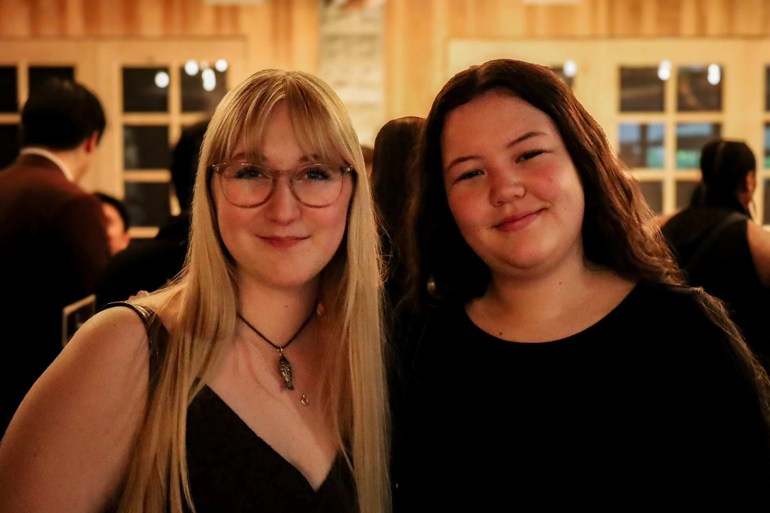 Two women with smiling faces standing close together indoors. The woman on the left has long blonde hair, glasses, and is wearing a black top. The woman on the right has long dark hair and is dressed in a black top. The background reveals a busy, war