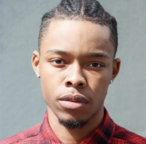 Close-up portrait of a young man with braided hair and earrings, wearing a red plaid shirt against a plain backdrop.