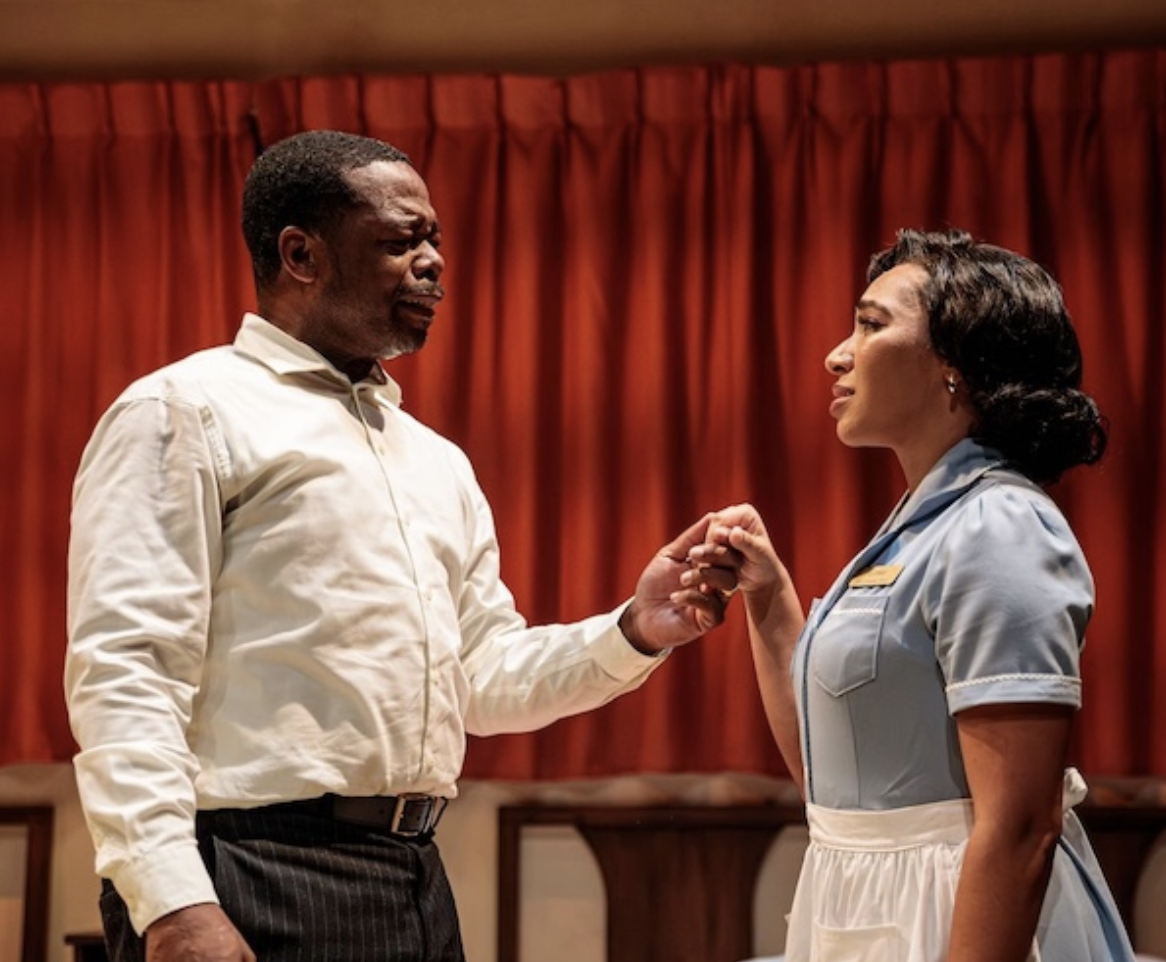 A man and a woman holding hands and looking at each other on a stage with red curtains in the background.