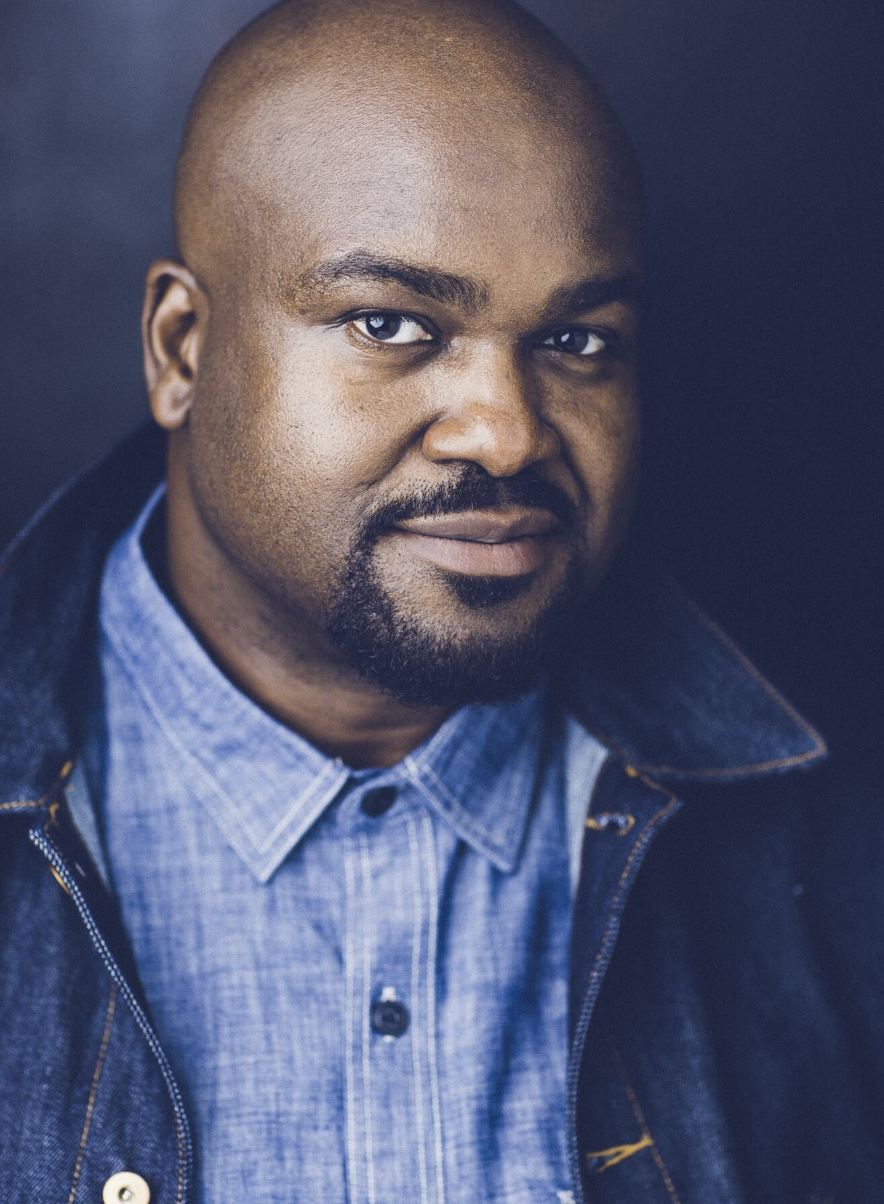 A portrait of a Black man with a shaved head, dark beard, and mustache, wearing a blue collared shirt and a dark denim jacket, against a dark background.