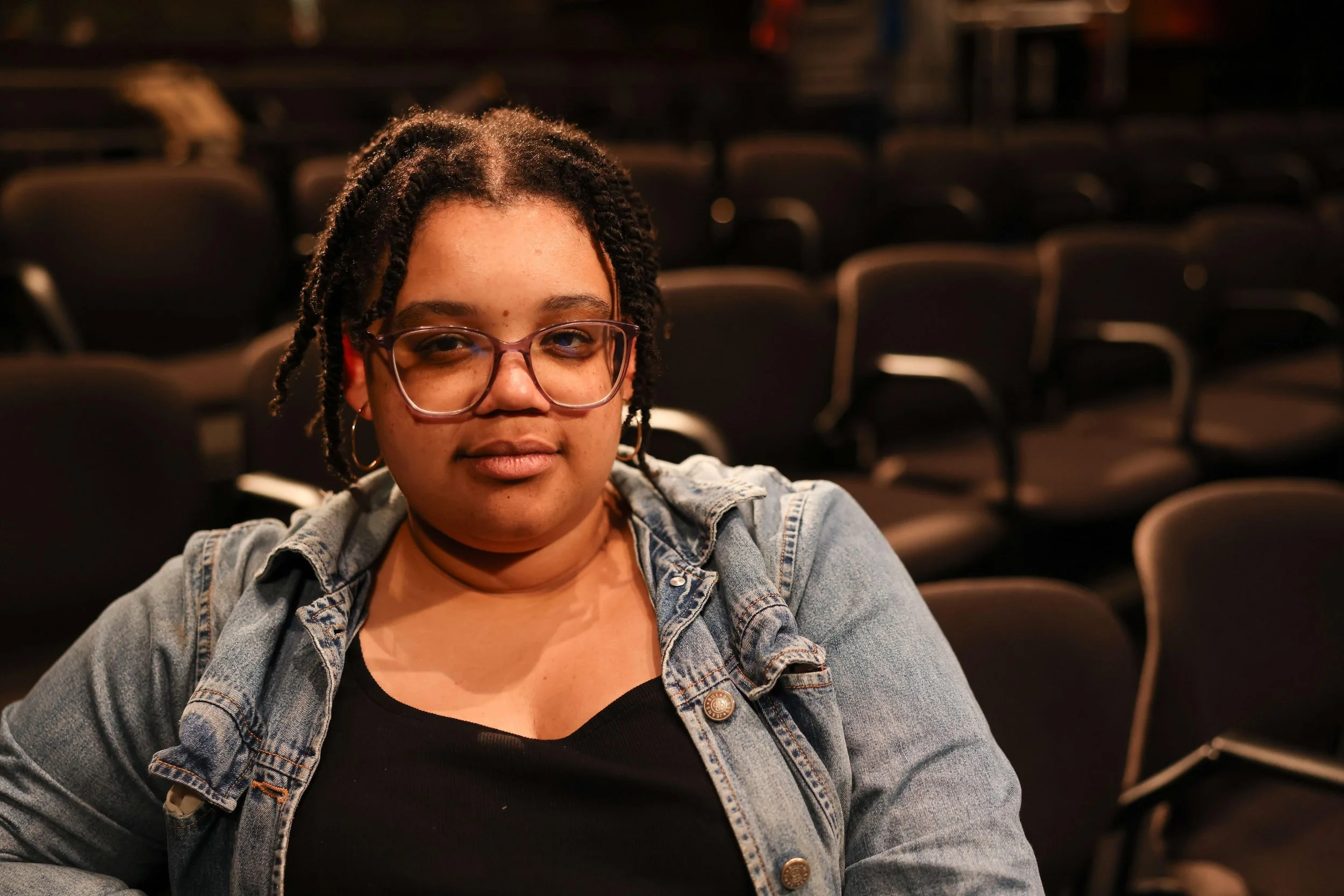 A young woman with glasses and styled hair sitting in an auditorium with empty chairs in the background.