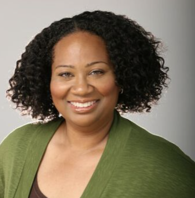 Smiling woman with curly black hair wearing a green jacket and a brown top, posing against a plain gray background.