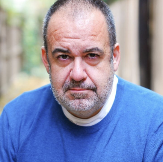 Close-up of a middle-aged man with a serious expression, wearing a blue shirt, outdoors with a blurred natural background.