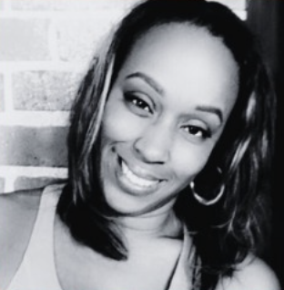A smiling woman with straight hair, hoop earrings, and a tank top, standing in front of a brick wall.