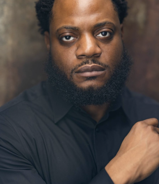 Close-up portrait of a man with dark skin, short curly hair, and a beard, wearing a black shirt, looking directly at the camera with a serious expression.