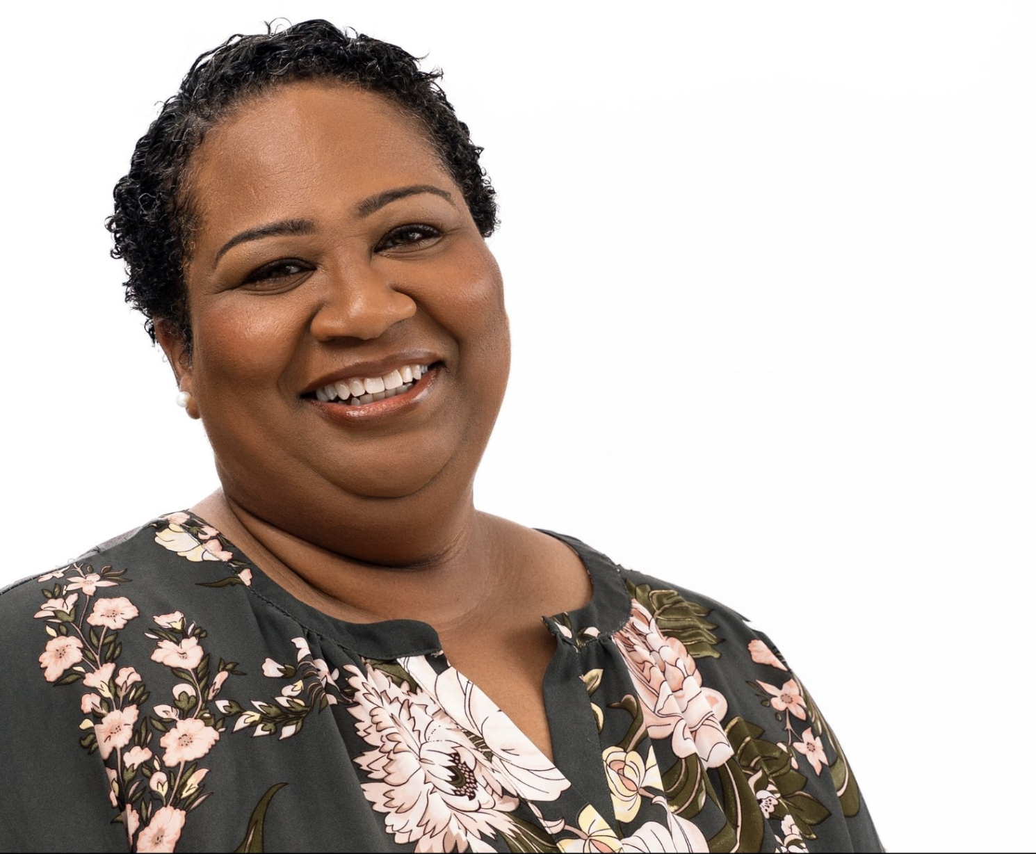 A woman with short curly hair, smiling, wearing makeup, pearl earrings, and a floral blouse against a white background.