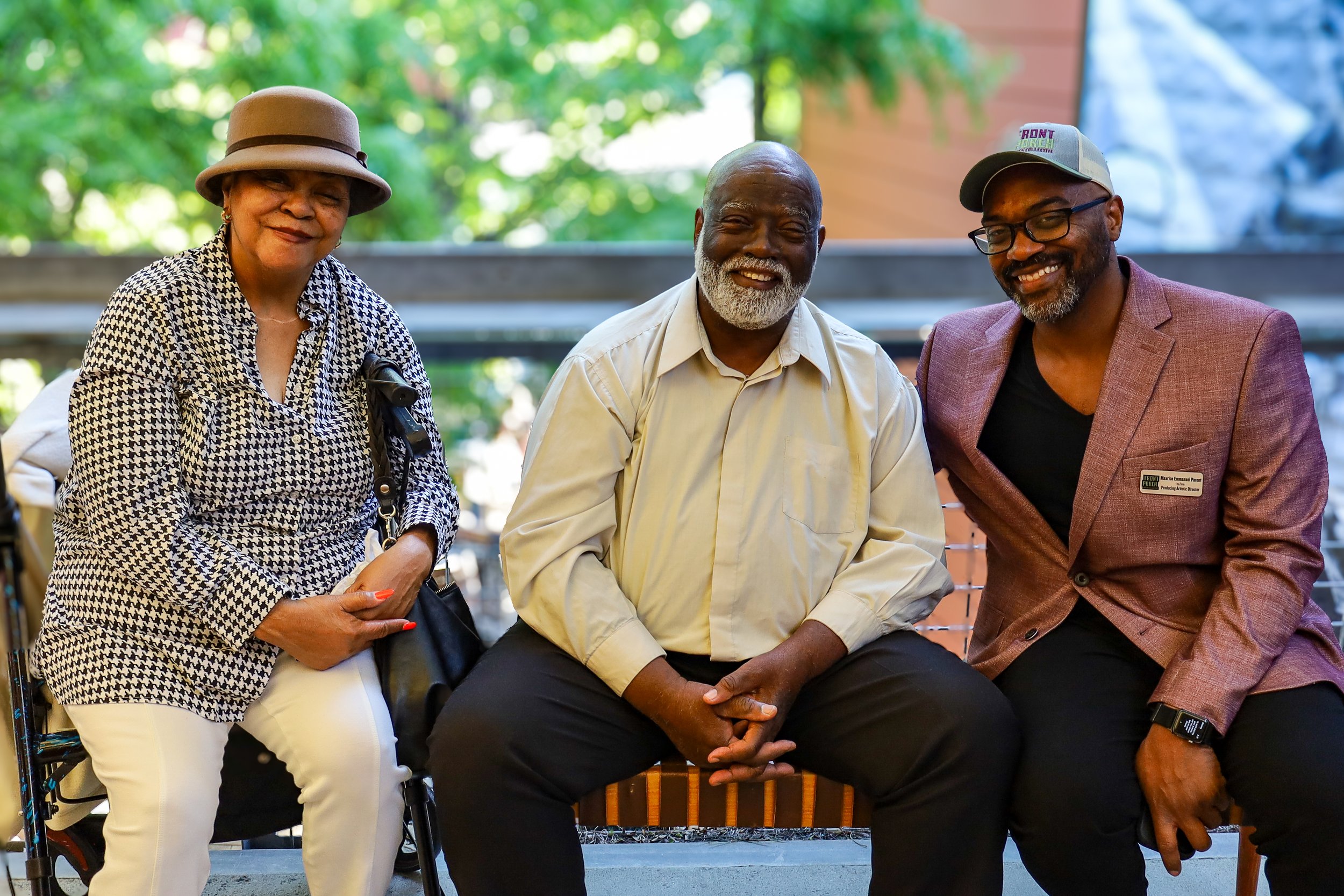 Three people sitting outdoors on a bench smiling, with greenery in the background. The woman on the left wears a hat and patterned blouse, the man in the center has a beard and wears a light shirt, and the man on the right wears glasses, a hat, and a