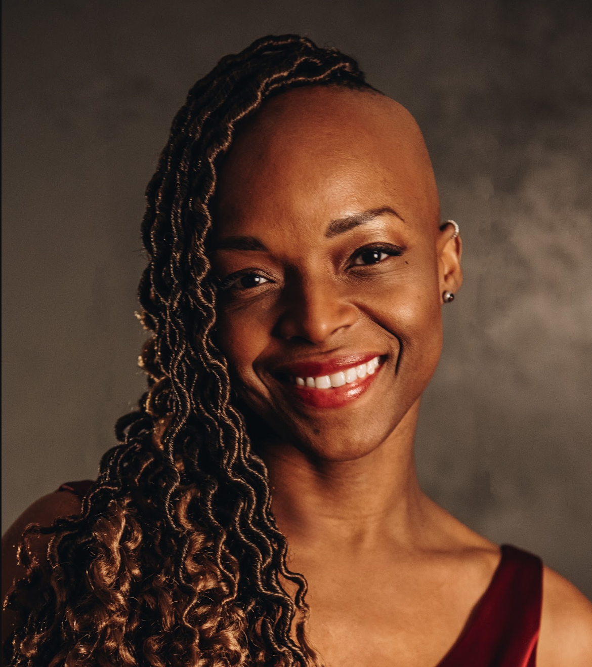 Portrait of a smiling woman with a shaved head and long, twisted hair on one side, wearing a red dress and earrings.